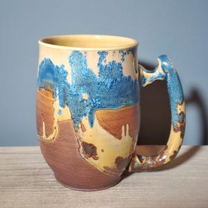 May include: A handmade ceramic mug with a unique, textured design. The mug features a combination of colors, including brown, tan, and blue, with a distinctive handle. The mug is approximately 5 inches tall and is suitable for hot or cold beverages.