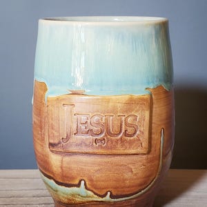 May include: A handmade ceramic mug with a light blue glaze on the top half and a brown, textured lower half. The word "JESUS" is embossed on the front. The mug has a rustic, artisanal appearance.