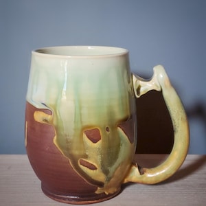May include: Handmade ceramic mug with a unique design. The mug features a brown base, transitioning to green and cream hues towards the top. The handle is a curved, organic shape. This stoneware mug is perfect for coffee or tea.