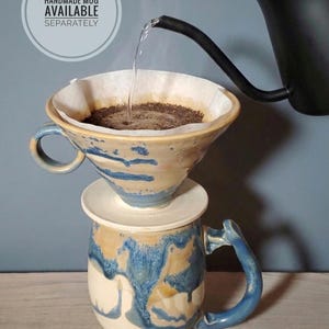 May include: A handmade ceramic mug with blue and tan glaze is shown with a pour-over coffee setup. A paper filter holds coffee grounds as hot water is poured from a black gooseneck kettle. The mug has a matching lid and handle. Text on the image reads "HANDMADE MUG AVAILABLE SEPARATELY."