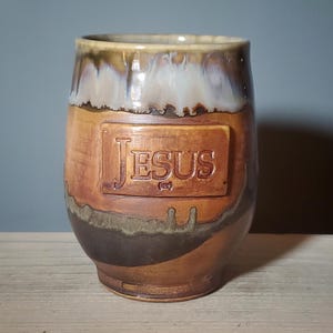 May include: A handmade ceramic tumbler with a brown and tan glaze. The word "JESUS" is embossed on a rectangular plaque on the front of the tumbler. The tumbler has a wide mouth and a rounded base.