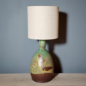 May include: A table lamp featuring a cylindrical off-white fabric lampshade atop a ceramic base. The base has a rounded, bottle-like shape with a glazed finish in shades of green, brown, and tan, transitioning to a solid dark brown bottom.