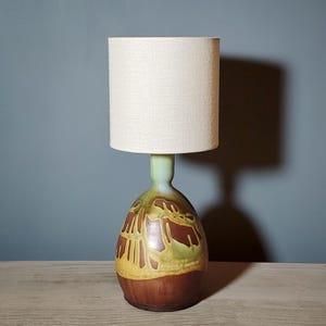 May include: A table lamp featuring a cylindrical off-white fabric shade atop a bulbous ceramic base. The base displays a multi-colored glaze with sections of deep brown, mustard yellow, and pale green in a textured, abstract pattern.