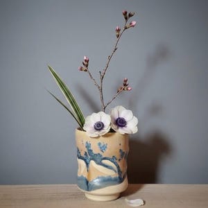 May include: A ceramic vase with a beige and blue design holds white anemones and a budding branch. A green leaf adds contrast. The vase sits on a wooden surface against a gray background, creating a serene floral arrangement.