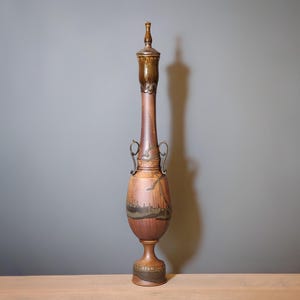 May include: A tall, decorative vase with a slender neck and a bulbous base. The vase is primarily brown with darker accents and metal handles. The top has a decorative finial. The vase is approximately 30 inches tall.
