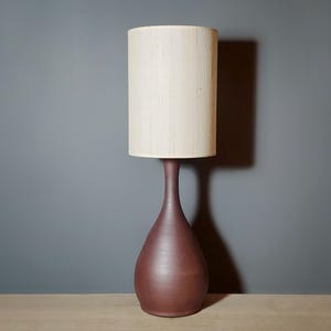 May include: A tall, burgundy-colored table lamp with a cream-colored lampshade. The lamp has a teardrop-shaped base and a slender neck. The lampshade is cylindrical. The lamp is set against a gray wall and a wooden surface.