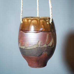 May include: A hanging ceramic planter with a brown and reddish-brown glaze. The top section is a glossy, dark brown, transitioning to a lighter reddish-brown and then a darker brown at the base. The planter is suspended by three beige ropes.