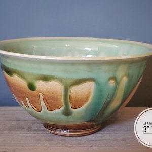 May include: A handmade ceramic bowl with a light green interior and exterior, featuring brown and green dripped glaze. The bowl is approximately 3 inches tall and has a wide, rounded shape, suitable for serving or display.