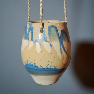 May include: A hanging ceramic planter with a cream-colored base and a blue and cream glaze. The planter is suspended by three woven rope cords. The blue glaze drips down the sides of the planter, creating a unique pattern. The planter is designed for indoor or outdoor use.