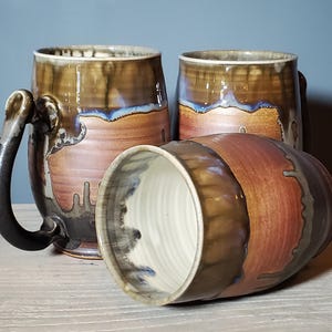 May include: Three handmade ceramic mugs with a rustic design. The mugs feature a brown and reddish-brown glaze with blue accents. One mug is lying on its side, revealing a white interior.