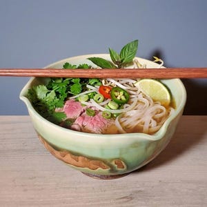 May include: A bowl of pho, a Vietnamese noodle soup, with chopsticks resting on top. The soup contains noodles, meat, herbs, and a lime wedge. The bowl is light green with brown accents.