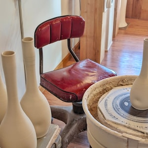 May include: A pottery studio scene featuring several unfinished, slender, off-white vases. A red leather chair sits in the background. One vase is on a pottery wheel. The vases are tall and have a narrow neck and rounded body.