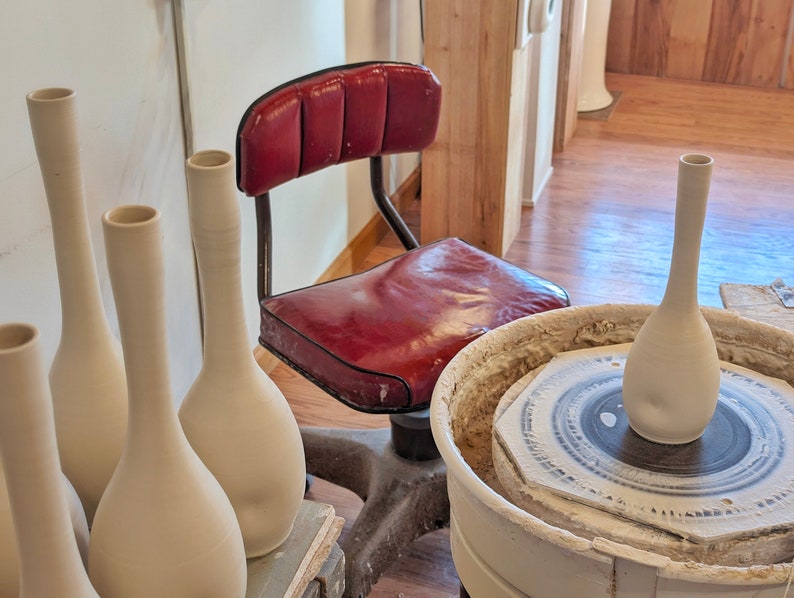 May include: A pottery studio scene featuring several unfinished, slender, off-white vases. A red leather chair sits in the background. One vase is on a pottery wheel. The vases are tall and have a narrow neck and rounded body.