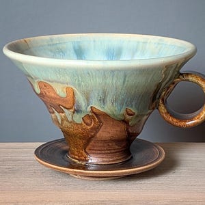 May include: Handmade ceramic pour-over coffee maker with a matching saucer. The coffee maker features a light blue and brown glaze with a circular handle. The design is a cone shape, perfect for brewing coffee. The saucer is a dark brown color.