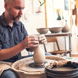 May include: A potter shapes a clay vase on a spinning pottery wheel. The artisan's hands are covered in wet clay as they mold the vessel. Other finished pottery pieces are visible in the background, showcasing the craft.