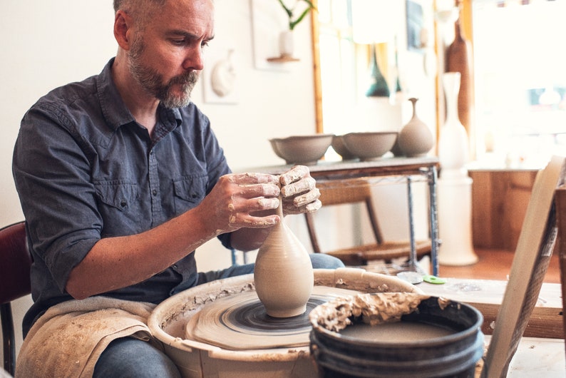 May include: A potter shapes a clay vase on a spinning pottery wheel. The artisan's hands are covered in wet clay as they mold the vessel. Other finished pottery pieces are visible in the background, showcasing the craft.
