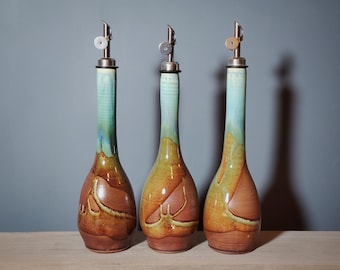 May include: Three ceramic oil bottles with stainless steel spouts. The bottles have a long neck with a turquoise glaze that fades into a brown and yellow body. The bottles are on a wooden surface.