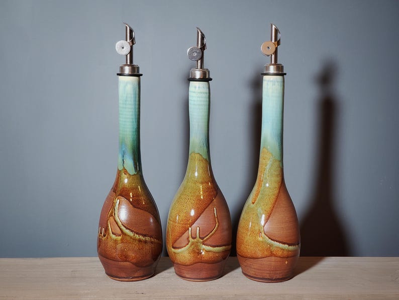 May include: Three ceramic oil bottles with stainless steel spouts. The bottles have a long neck with a turquoise glaze that fades into a brown and yellow body. The bottles are on a wooden surface.