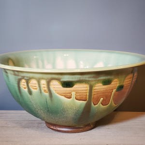 May include: A large, handmade ceramic bowl with a wide rim. The bowl features a green glaze with brown and dark green decorative elements. The bowl is approximately 12 inches in diameter and sits on a small, round base.