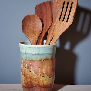 May include: A ceramic utensil holder with a brown and orange glaze, holding wooden spoons and spatulas. The holder has a light blue rim and a dripped glaze design. The wooden utensils include a spoon, a spatula, and a slotted spatula.