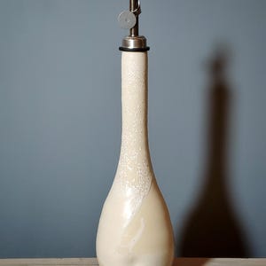 May include: A tall, slender ceramic oil cruet with a cream-colored glaze featuring a textured, speckled finish on the upper neck. The bottle is topped with a silver-toned metal pourer spout that includes a small, circular hinged lid mechanism.