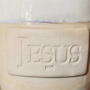 May include: A cream-colored ceramic jar with a white top. The word "JESUS" is embossed on a rectangular panel on the jar's body. The jar has a rustic, handmade appearance.