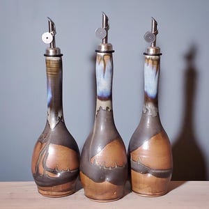 May include: Three ceramic oil bottles with stainless steel spouts. The bottles have a brown and gray color scheme with blue accents. The bottles are displayed on a wooden surface against a blue-gray background.