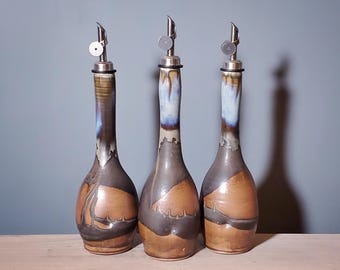 May include: Three ceramic oil bottles with stainless steel spouts. The bottles have a brown and gray color scheme with blue accents. The bottles are displayed on a wooden surface against a blue-gray background.