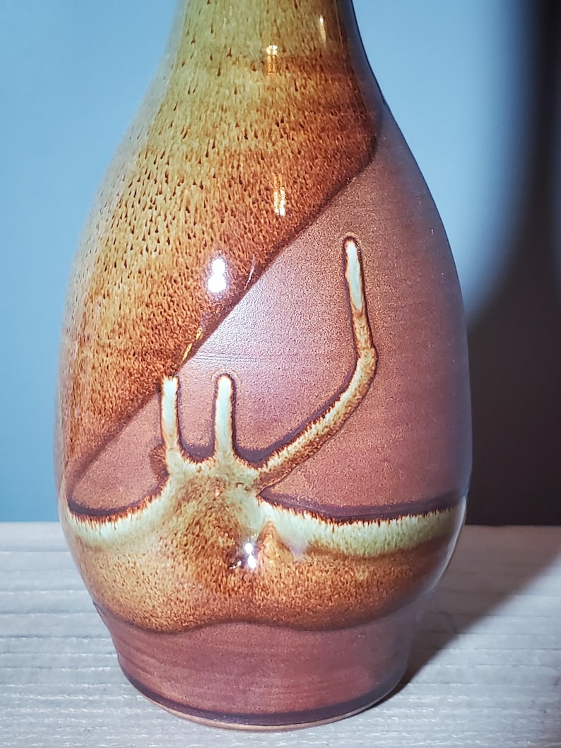 May include: A handcrafted ceramic vase with a unique design. The vase features a blend of brown, tan, and reddish-brown hues. The surface has a textured appearance with a distinctive abstract pattern in lighter tones.