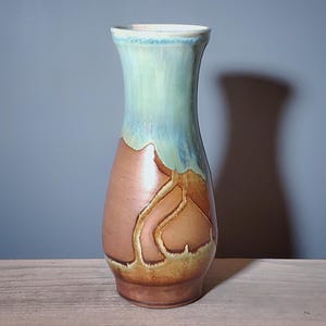 May include: A handcrafted ceramic vase with a unique design. The vase features a light blue and green upper section, transitioning to a brown and tan lower portion with an abstract pattern. The vase is displayed on a wooden surface.