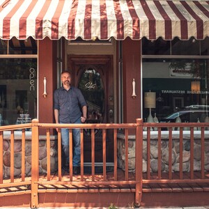 May include: Exterior shot of a small business with a red and white striped awning. A man stands in the doorway. The building has a red facade, a wooden porch, and a stone base. The windows display lamps and decorative items.