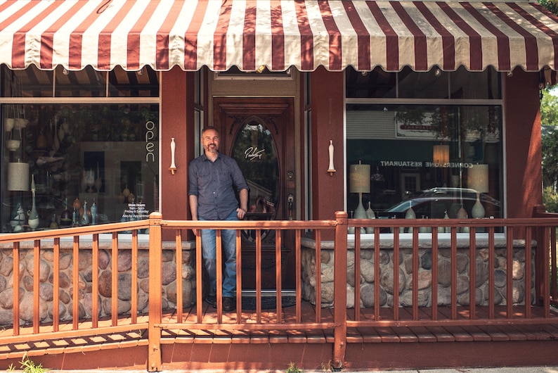 May include: Exterior shot of a small business with a red and white striped awning. A man stands in the doorway. The building has a red facade, a wooden porch, and a stone base. The windows display lamps and decorative items.