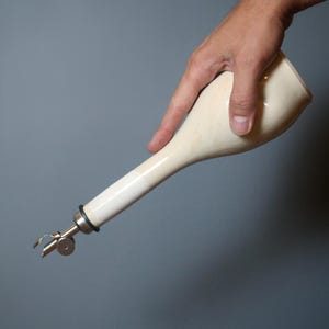 May include: Cream-colored ceramic oil bottle with a long, slender spout and a metal pourer. The bottle is held by a hand against a gray background. The pourer has a small, hinged spout for controlled pouring.