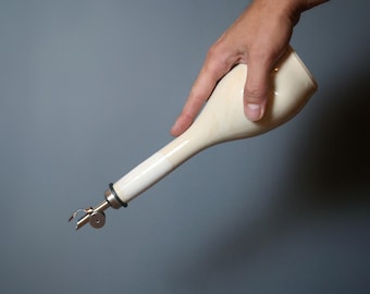 May include: Cream-colored ceramic oil bottle with a long, slender spout and a metal pourer. The bottle is held by a hand against a gray background. The pourer has a small, hinged spout for controlled pouring.