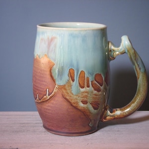 May include: A handmade ceramic mug with a unique design. The mug features a blend of colors, including light blue, brown, and orange. The handle is a continuation of the mug's design. The mug is a perfect size for coffee or tea.