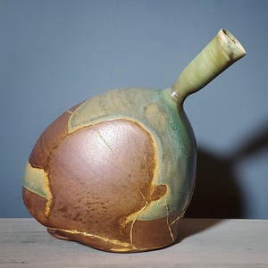 May include: A handcrafted ceramic vase with a bulbous body and a slender, angled spout. The vase features a blend of earthy tones, including brown, green, and yellow, with visible crack patterns adding to its unique aesthetic. The vase is a decorative item.