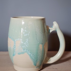 May include: A handmade ceramic mug with a unique handle design. The mug features a light blue glaze that drips over a creamy white base, creating a textured, artistic effect. The mug is suitable for hot or cold beverages.