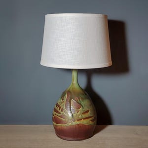 May include: A ceramic table lamp with a white linen shade. The lamp base is a bulbous shape with a green, brown, and red glaze. The base features a decorative relief design. The lamp is on a wooden surface.