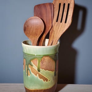 May include: A ceramic utensil holder with a green and brown glaze, holding wooden spoons and spatulas. The holder has a cylindrical shape with a light green rim and a brown base. The wooden utensils are various shapes and sizes.