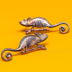 Puede incluir: Dos broches de camaleón plateados sobre un fondo amarillo brillante. Los broches tienen escamas texturizadas y colas rizadas. Un broche está posado en una pequeña rama. Los broches son detallados.