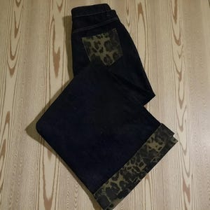 Puede incluir: Vaqueros de mezclilla azul oscuro con un bolsillo y un detalle de puño con estampado de leopardo. Los vaqueros están doblados, mostrando el estampado animal contrastante con la tela azul oscuro. Estilo clásico.