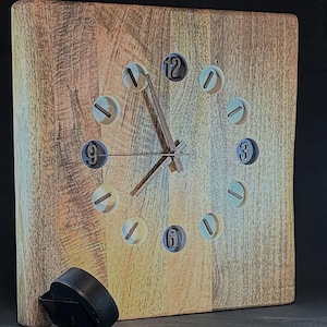May include: Square wooden clock with a light brown finish. The clock face features screw-shaped hour markers and clock hands. A black object rests on the lower left side of the clock. The clock is a decorative item.