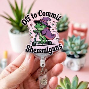 May include: A retractable badge reel featuring a cartoon frog on a scooter, dressed in a witch hat and cape. The text says "Off to Commit Shenanigans". The design is set against a pink backdrop.