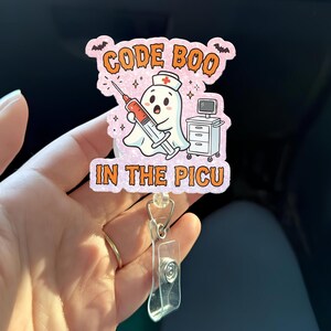 May include: A novelty badge reel featuring a cartoon ghost nurse holding a syringe with red liquid. The text "CODE BOO" and "IN THE PICU" are displayed in orange. The badge reel has a clear plastic clip and a retractable cord.