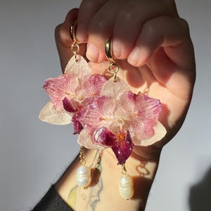 Peut inclure: Boucles d'oreilles faites à la main avec des fleurs d'orchidées roses et blanches pressées. Les boucles d'oreilles ont des cerceaux et des chaînes dorés, avec une seule perle blanche sur chaque boucle. Une main tient les boucles d'oreilles.