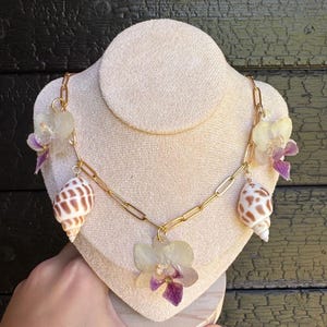 Peut inclure: Un collier à chaîne dorée au design unique. Le collier présente trois coquillages et trois grappes de fleurs séchées avec des accents violets. Le collier est exposé sur un buste crème.