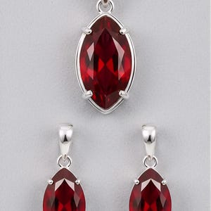 Puede incluir: Un conjunto de joyas que incluye un collar con colgante y pendientes a juego. El conjunto presenta una piedra preciosa roja oscura con corte marquesa engastada en metal plateado. El colgante y los pendientes tienen un diseño similar.