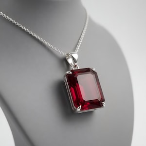 Puede incluir: Un collar de plata con un gran colgante rectangular de piedra preciosa rojo oscuro. El colgante está engastado en un marco de plata y cuelga de una delicada cadena de plata. La piedra preciosa mide aproximadamente 2.5 cm de largo y 2 cm de ancho.