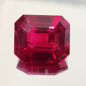 Puede incluir: Una gema de rubí tallada en forma rectangular facetada. La gema es de color rojo intenso y saturado, con múltiples facetas que reflejan la luz. La piedra preciosa se muestra sobre una superficie reflectante.