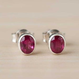 Puede incluir: Un par de pendientes de botón con piedras preciosas rojas oscuras de forma ovalada engastadas en plata. Los pendientes se exhiben sobre una superficie de madera clara, mostrando su apariencia pulida y su color vibrante.